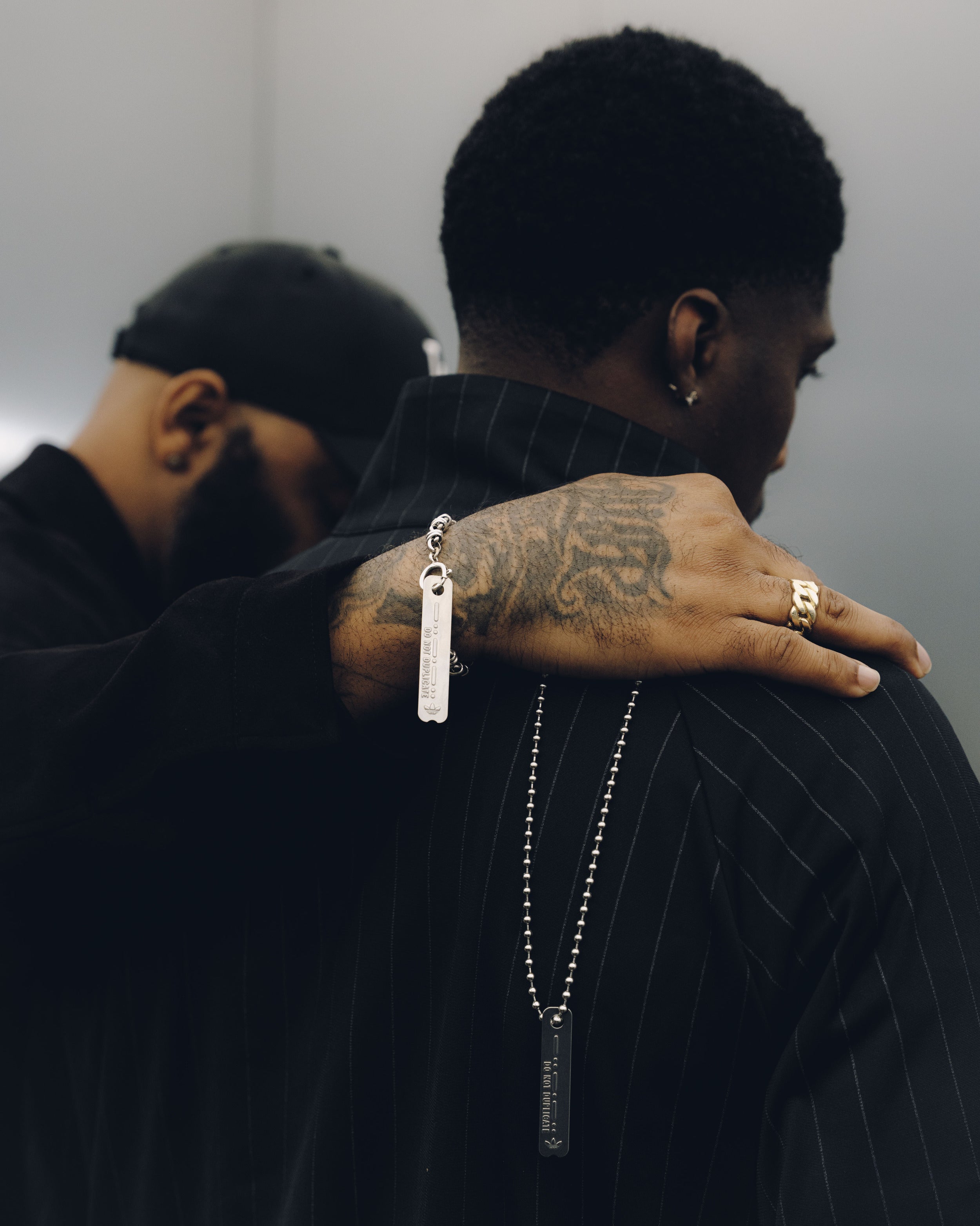 Close-up of two men in dark streetwear, one with tattooed hand and layered silver necklaces.
