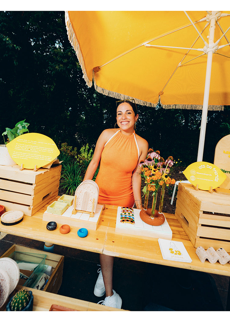 Woman in orange dress at outdoor market booth with handmade crafts and yellow umbrella