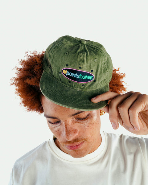Man wearing olive green embroidered cap with all points bulletin logo and white t-shirt