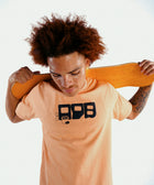 Man in peach streetwear t-shirt with graphic print holding skateboard behind shoulders