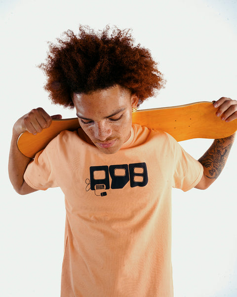 Man in peach streetwear t-shirt with graphic print holding skateboard behind shoulders