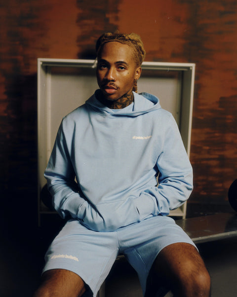 Man wearing a light blue hoodie and shorts set with 'all points bulletin.' text, seated indoors