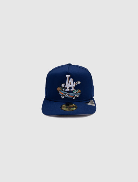 Blue fitted baseball cap with LA logo and cartoon skateboard graphic, streetwear style.