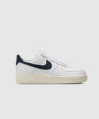 White low-top sneaker with dark blue swoosh, cream sole, and perforated toe on white background