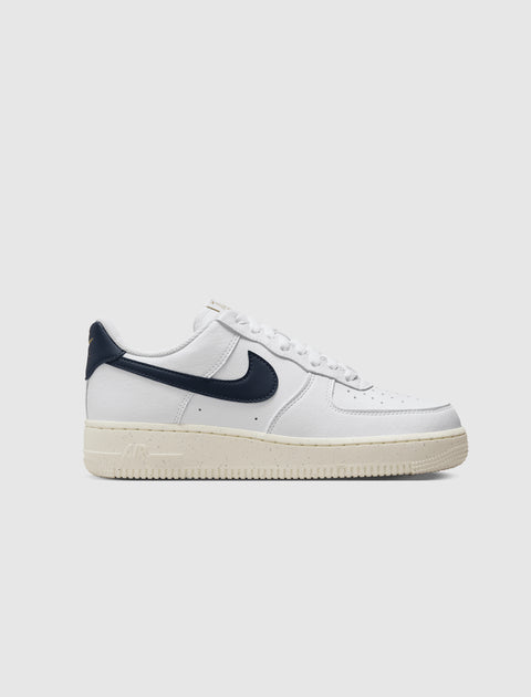 White low-top sneaker with dark blue swoosh, cream sole, and perforated toe on white background