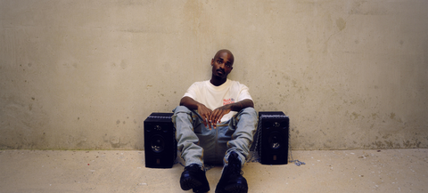 Man in streetwear sits against a concrete wall with speakers, wearing a white graphic tee and jeans.