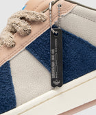 Close-up of a textured sneaker with beige, blue, and cream panels, chunky laces, and a metal tag.