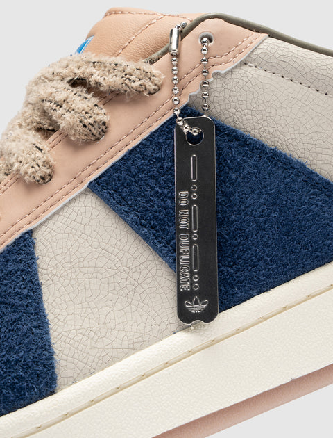 Close-up of a textured sneaker with beige, blue, and cream panels, chunky laces, and a metal tag.