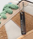 Closeup of a streetwear sneaker with textured brown panels, cracked white leather, fuzzy green laces, and metal tag reading 'Do Not Duplicate'.
