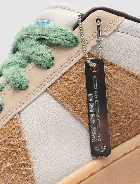 Closeup of a streetwear sneaker with textured brown panels, cracked white leather, fuzzy green laces, and metal tag reading 'Do Not Duplicate'.