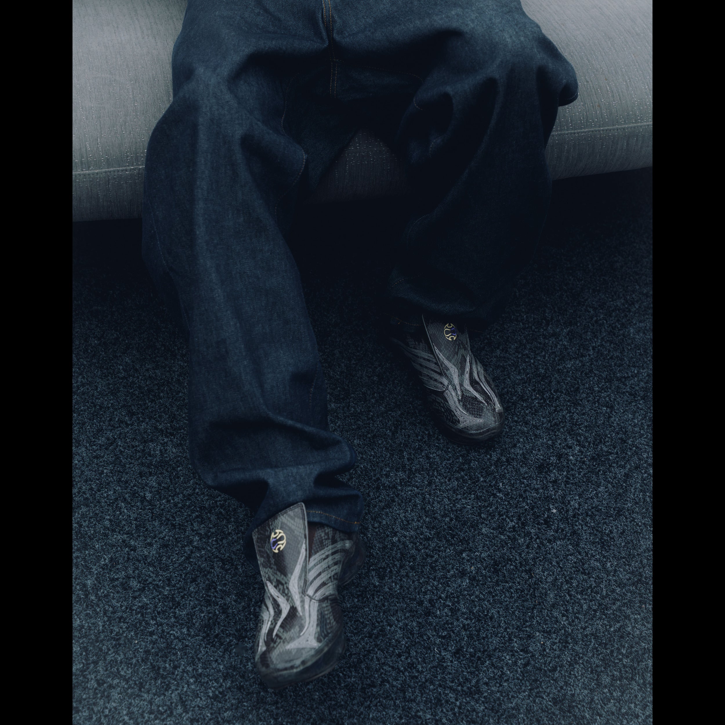 Person wearing baggy dark denim jeans and patterned sneakers, seated on a modern sofa.