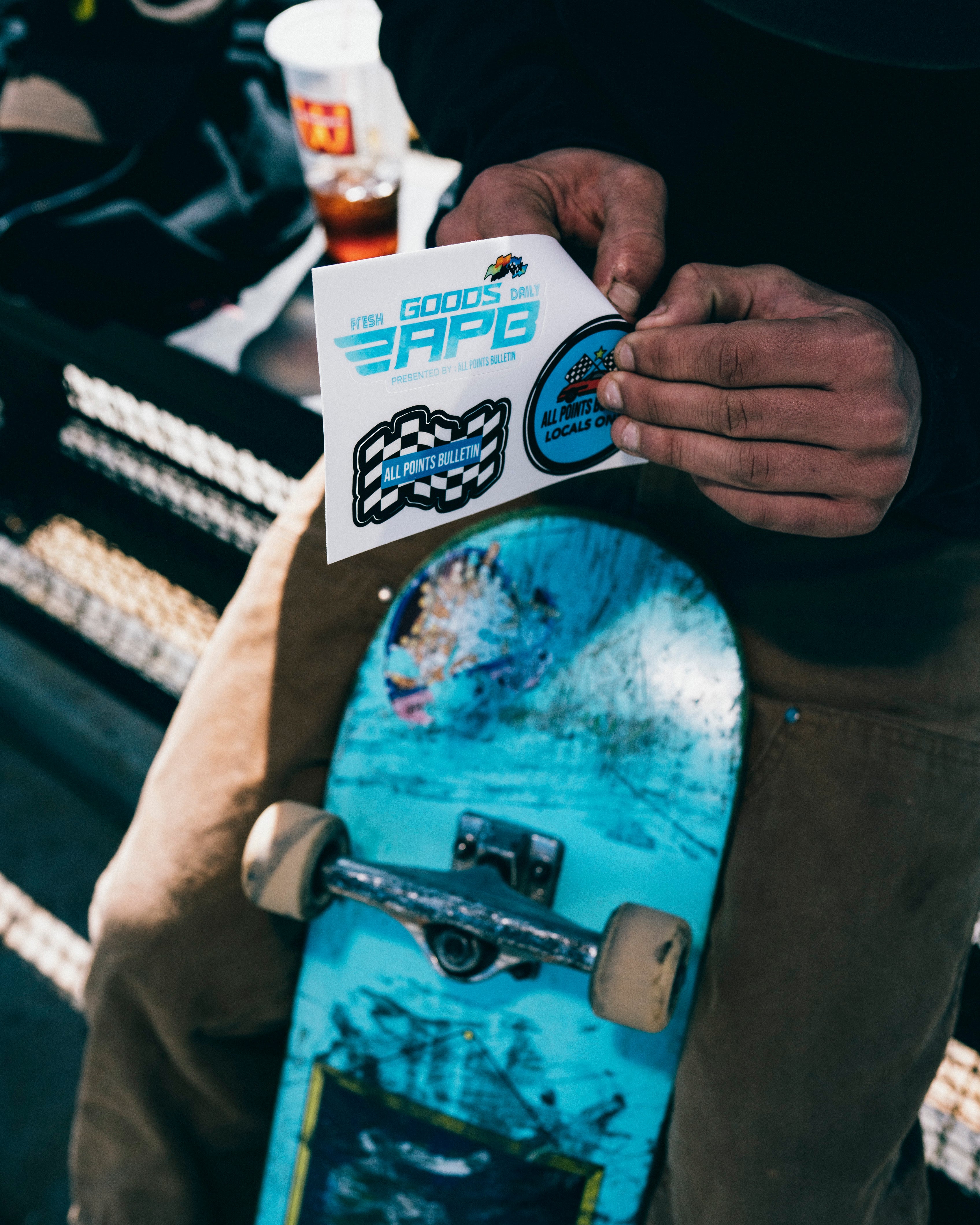 Person holding streetwear stickers above a blue skateboard, casual urban setting