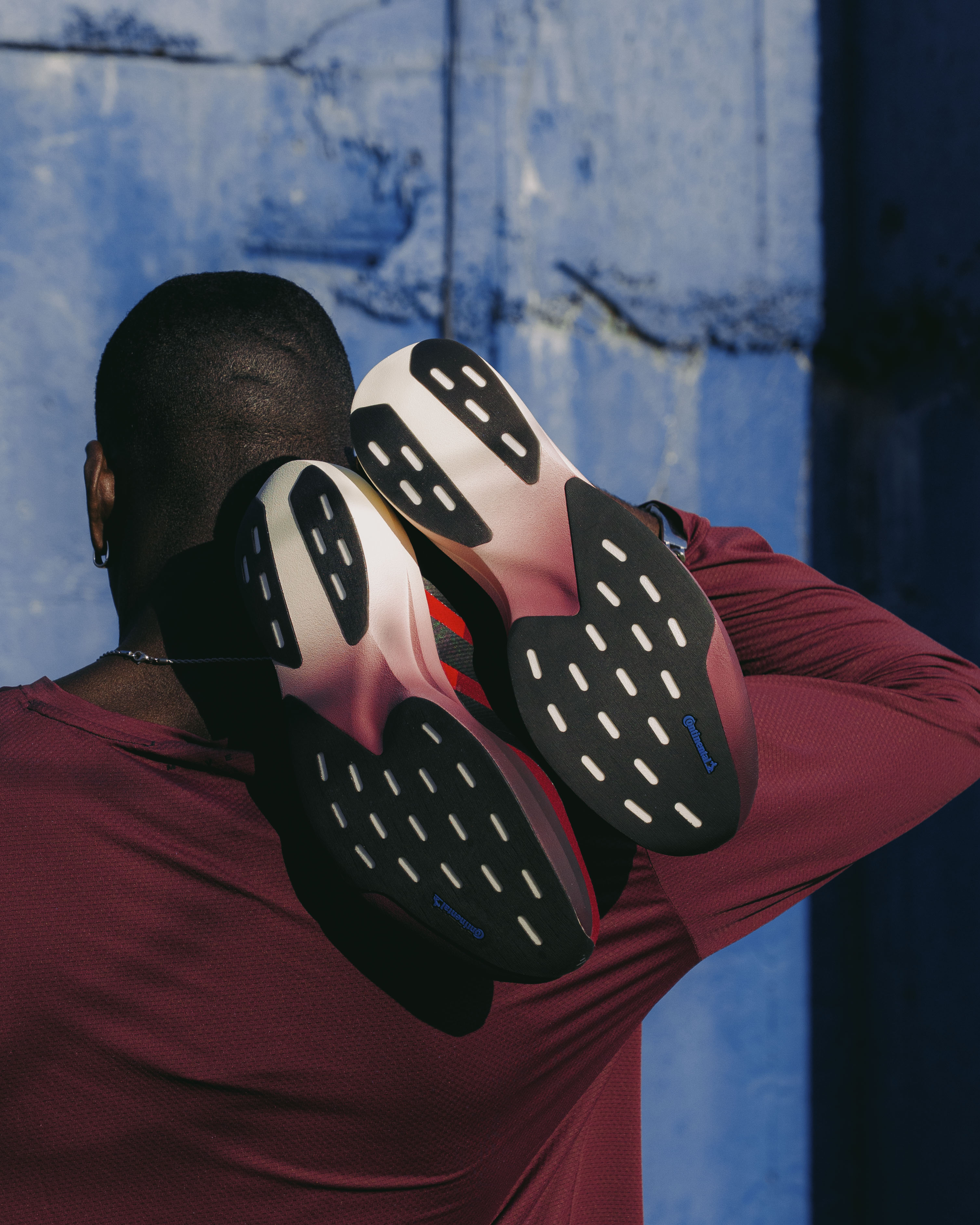 Man in burgundy streetwear holding sneakers over shoulder against blue wall