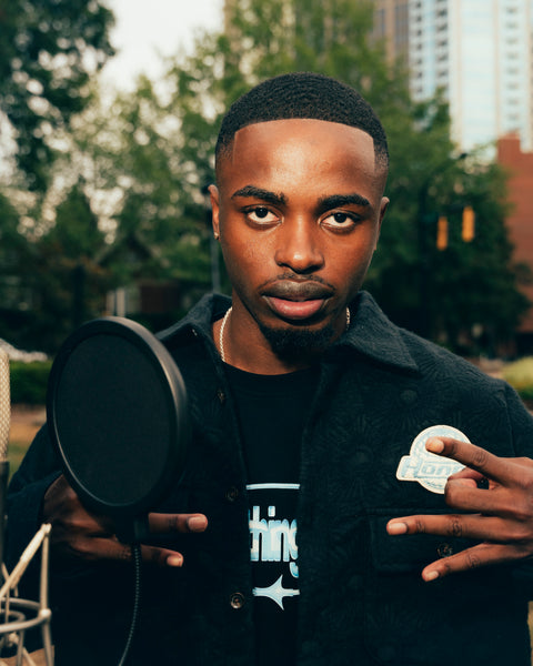 Man in streetwear jacket and graphic tee posing outdoors by a microphone in the city
