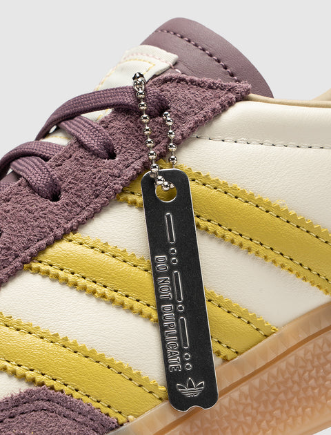 Close-up of sneaker with purple suede, yellow stripes, and metal tag reading 'DO NOT DUPLICATE'
