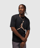 Man wearing black graphic streetwear t-shirt with large camo logo, gray pants, chain necklace