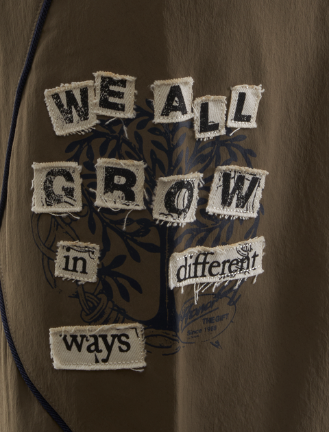Brown streetwear shirt with sewn patchwork text saying 'We all grow in different ways'.