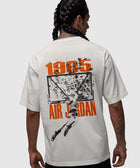Back view of a man wearing a white Air Jordan 1985 graphic t-shirt, featuring a basketball dunk illustration.