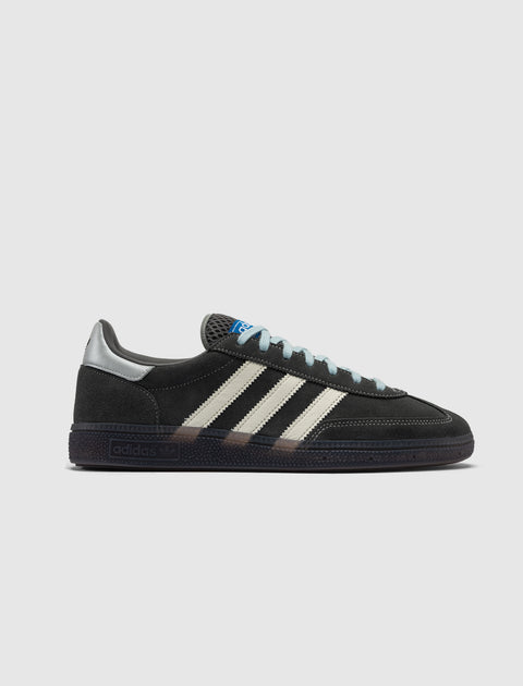 Black suede sneaker with white stripes, grey heel tab, blue label, and gum sole on white background