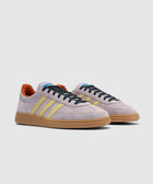 Lilac suede sneakers with yellow stripes, gum sole, and orange heel accents on white background