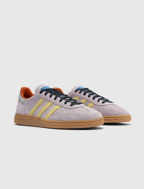 Lilac suede sneakers with yellow stripes, gum sole, and orange heel accents on white background