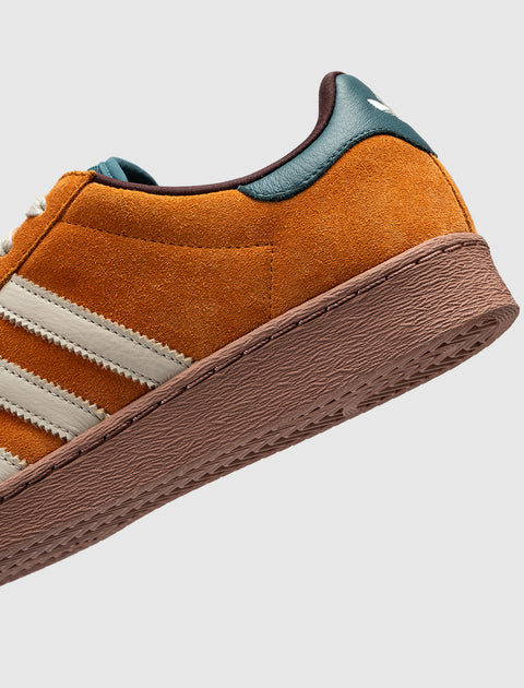 Close-up of brown suede sneaker with white stripes, teal heel tab, and textured sole
