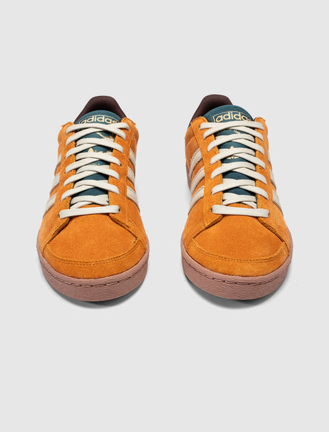 Pair of orange suede Adidas sneakers with cream stripes and rubber soles on a plain background