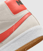 Close-up of light gray suede sneaker with orange leather heel, orange swoosh, and textured white sole.