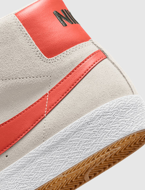 Close-up of light gray suede sneaker with orange leather heel, orange swoosh, and textured white sole.