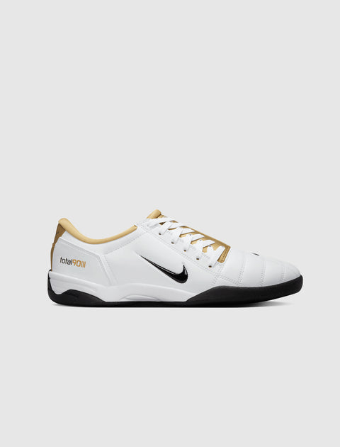 White and gold Nike sneaker with black sole and Total 90 branding, side view on gray background