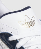 Close-up of a white sneaker with navy and green accents, gold logo, and white laces