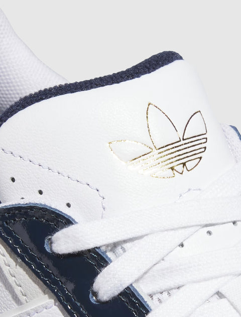 Close-up of a white sneaker with navy and green accents, gold logo, and white laces