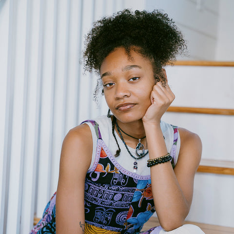 Woman with curly hair wearing a colorful patterned top and layered necklaces on stairs