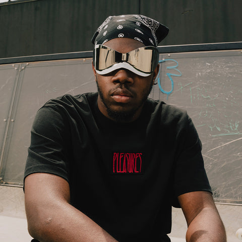 Man in streetwear with black bandana, futuristic sunglasses, and PLEASURES black t-shirt