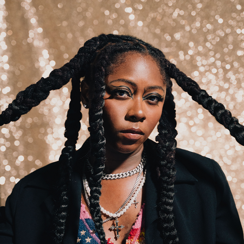 Fashionable woman with long braids, layered necklaces, and bold makeup in front of a glitter backdrop