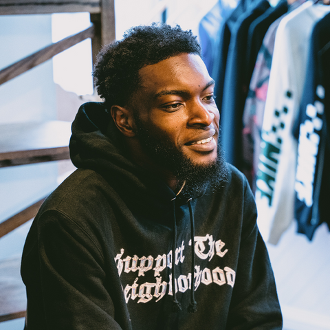 Smiling man in black streetwear hoodie with gothic text, apparel racks in background