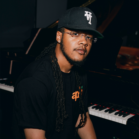 Man in black streetwear t-shirt and cap sitting at a piano in a modern setting