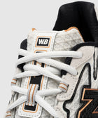 Close-up of white textured sneaker with black and gold accents, WB logo on tongue, streetwear style