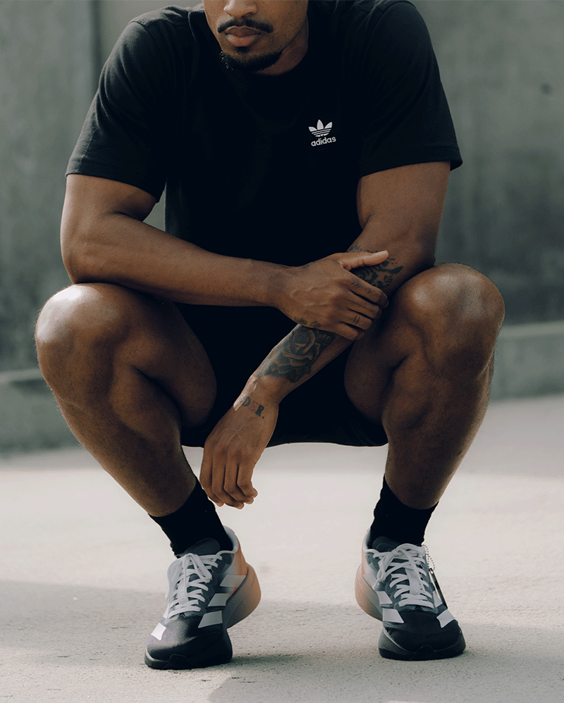 Man in black Adidas t-shirt and sneakers crouching outdoors, streetwear fashion style
