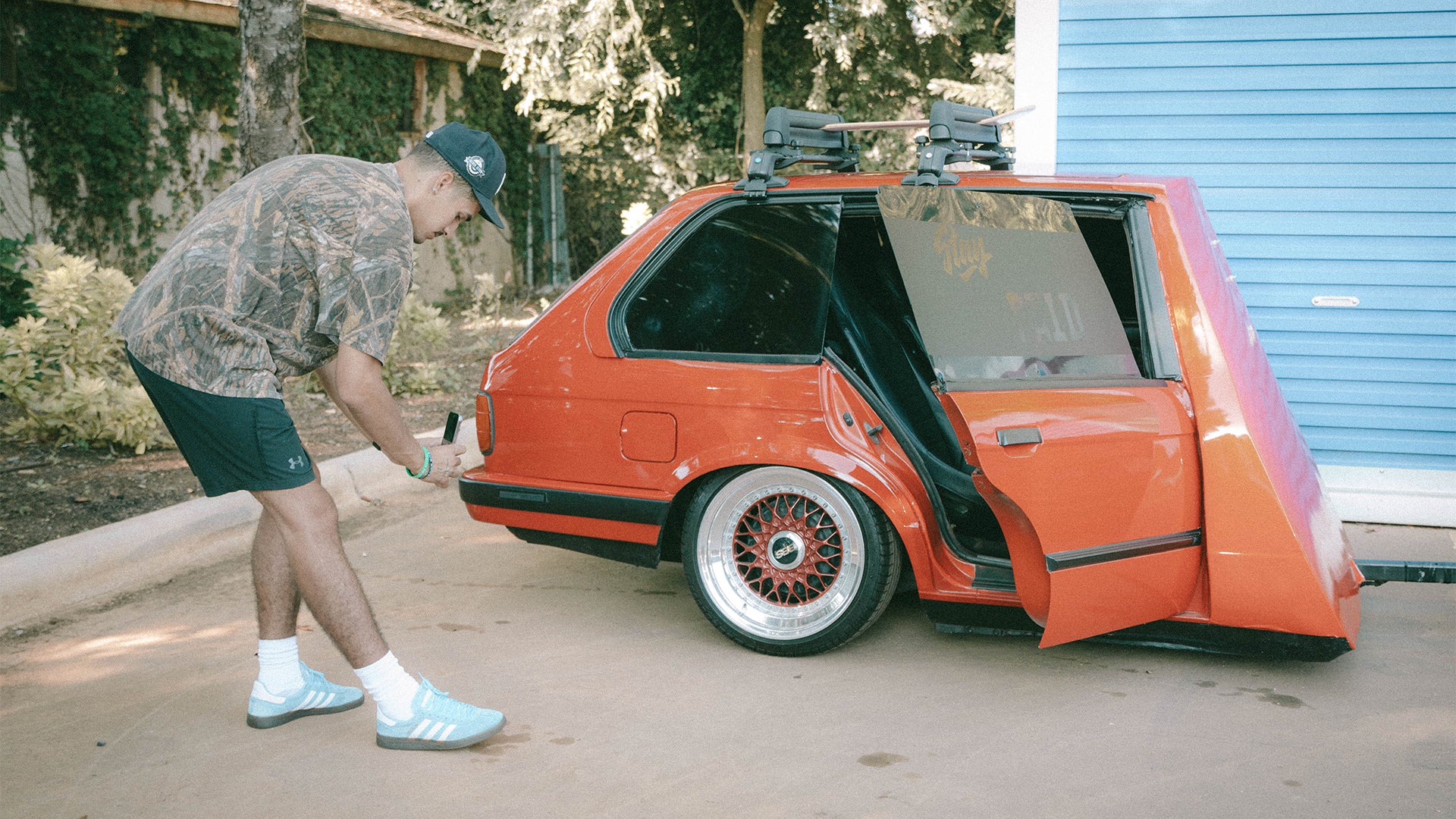 Man in streetwear next to custom red car with open door and sporty wheels outdoors