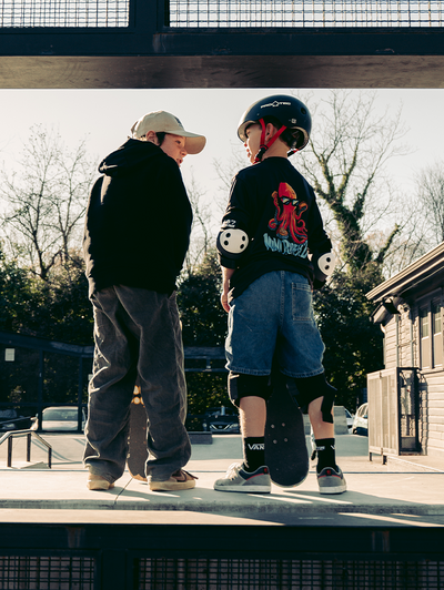Two kids at a skatepark, one with a helmet and skateboard, wearing streetwear