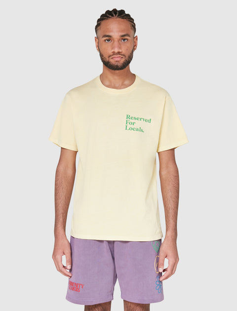 Man wearing a yellow t-shirt with 'Reserved For Locals' green text and lavender streetwear shorts.